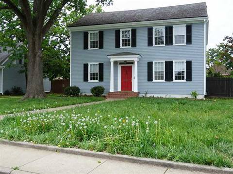 The front lawn of a two-storey house left long and going to seed, with dandelions through it and grass spilling over the sidewalk.